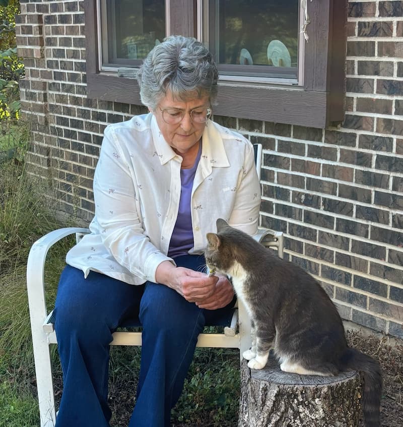 An older woman sits on a bench by a brick house, offering her hand to a gray and white cat sitting on a tree stump.
