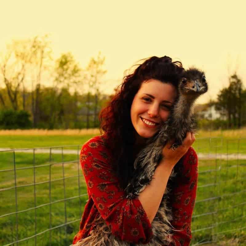 A woman with dark curly hair smiles while holding an emu close to her face, standing outside near a wire fence and grassy field.