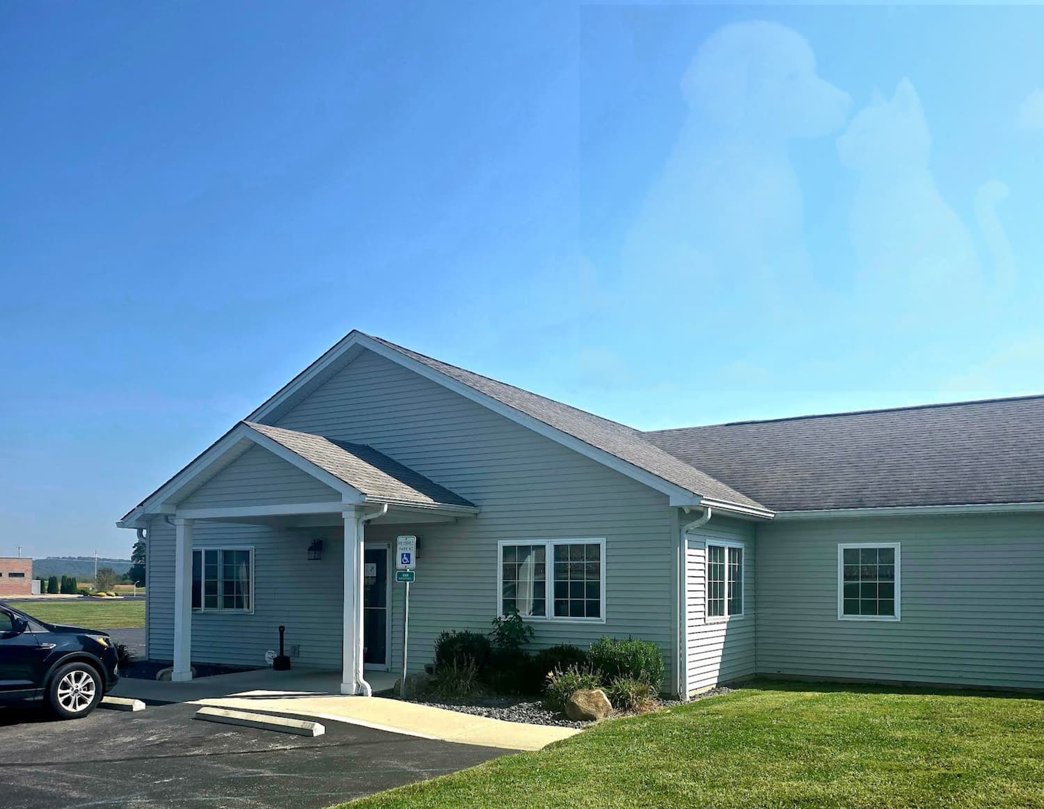Single-story building with gray siding, white trim, and accessible entrance; a car is parked outside and pet silhouettes are faintly visible in the sky.